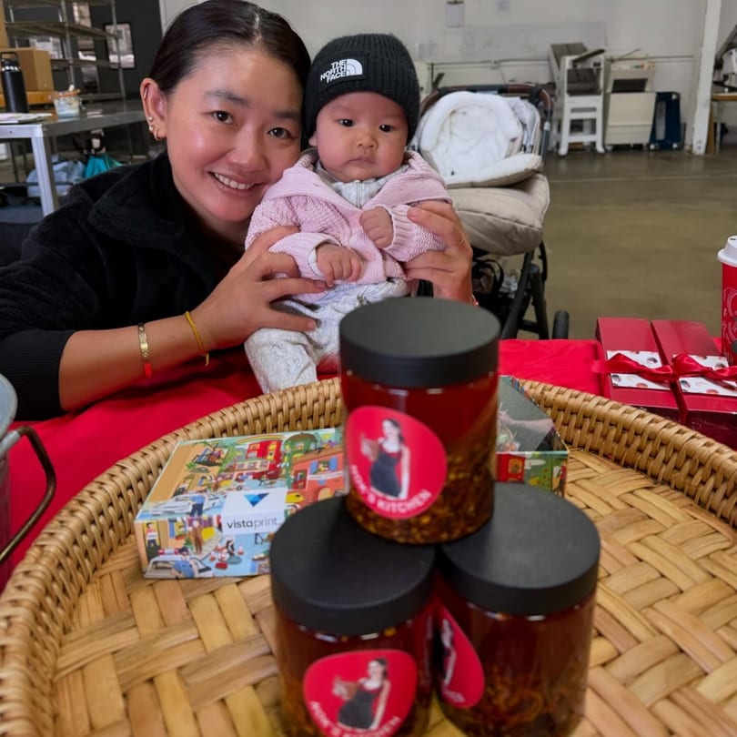 A woman holding a baby, with jars of Nok's Kitchen chili oil on a woven tray.